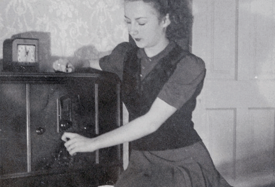 Black and white photo of woman sitting beside a 1940s radio, turning the knobs.