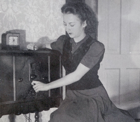 Black and white photo of woman sitting beside a 1940s radio, turning the knobs.